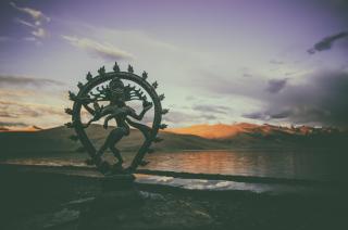 Shiva Nataraja at dusk on the shores of a placid lake, with mountains in the distance.