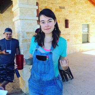 a young woman faces the camera, wearing overalls, with a coyp of "becoming" sticking out of her front pocket