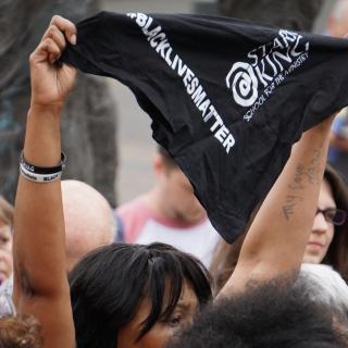 A Black woman raises a black bandana which reads "Black Lives Matter" in a crowd of people