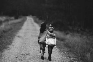 Black and white photo of two children walking away from the viewer on a straight dirt path, with brush and trees on either side. The taller child's arm is around the smaller child's shoulder. The smaller child is wearing a knit sweater and a striped knit hat with a pompom.