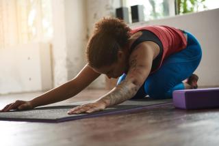 A Black woman in athletic clothing is in child's pose on a yoga mat: arms stretched forward, head bowing, kneeling back towards her heels.