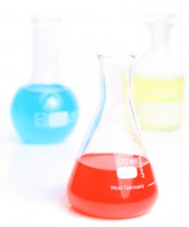 Three laboratory flasks sit on a white counter, each filled with a vividly colored liquid (sky blue, cherry red, and sunshine yellow)
