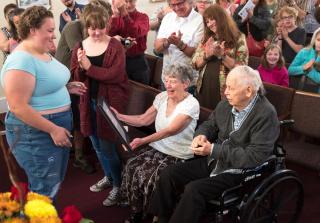 A standing youth presents an award to an sitting elderly couple, one in a wheelchair.