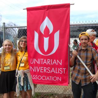 Youth wearing Standing on the Side of Love t-shirts stand by fence, holding UUA banner