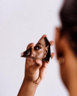 A Black man's hand is holding a mirror, in which we see a partial reflection of his face. Our point of view is from over his shoulder, so in the foreground we see a slightly out-of-focus portion of the back of his head.