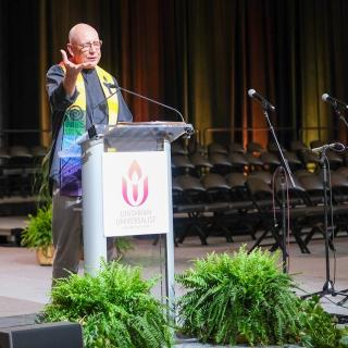 Rev. Jim Magaw stands at the podium of the General Assembly stage in Pittsburgh PA. Rev. Jim is a white man bald with black robes and a colorful stole. He speaks into the microphone with an intense and concentrated look on his face.