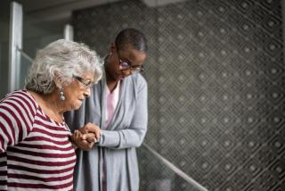 A Black woman with short-cropped hair helps an older woman walk down stairs.
