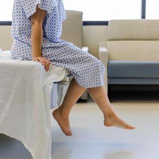 A person sits on a medical examination table, wearing a medical exam gown. Their head isn't visible but their feet dangle off the end and their arms hold the table tensely.