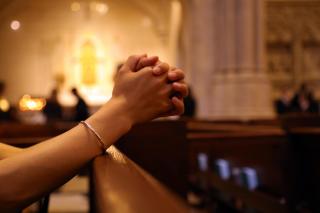 A person's hands are clasped in prayer in what appears to be a church pew.