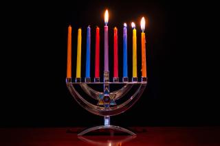 A silver menorah, against a dark background, holds colored candles. The first three candles and the shamash candle are lit.