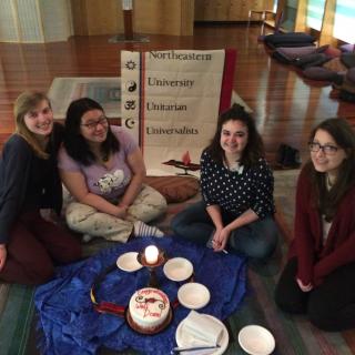 four young women sit in front of a sign for Northeastern Unitarian Universalists with an altar with a flaming chalice on it