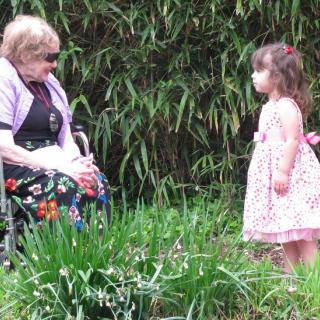 Woman in colorful skirt in wheelchair with young girl in pink dress amid green plants.