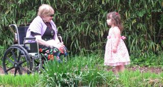 Woman in colorful skirt in wheelchair with young girl in pink dress amid green plants.