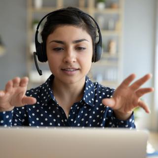 A woman wearing headphones sits at a laptop, gesturing with both hands at someone (not visible) on the screen.