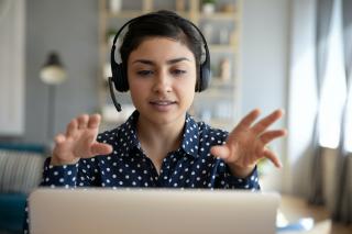 A woman wearing headphones sits at a laptop, gesturing with both hands at someone (not visible) on the screen.