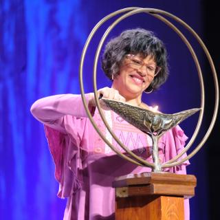 A woman lights the GA chalice. She has bangs and a chin-length bob, and her hair is dark while slightly graying. She wears a pink long sleeve blouse with fluttery sleeves and has pink rimmed glasses. She has a joyful look upon her face as she illuminates the chalice.