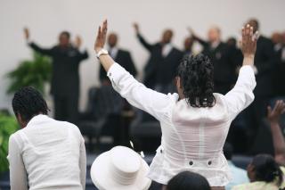 In the background, a group of Black people stand on a church chancel. In the foreground, from behind, a black person stands in their pew with arms raised in a "praise" position.