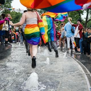 At a Pride celebration, people in various rainbow clothing joyfully gather while a few people splash through a public fountain.