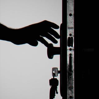 Silhouette of a hand reaching for a doorknob. The door key is still in the lock, and other keys dangle from the ring. Metal hardware gleams on the edge of the door.