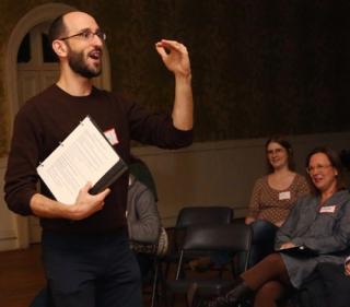 Holding a music binder and gesturing, Paul Vasile leads a group in singing