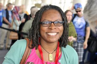 A woman with small square glasses, dreadlocks, and a flaming chalice necklace smiles.