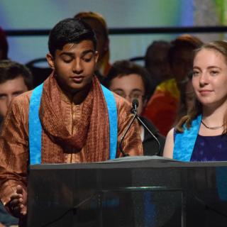 a crowd of young people are in the background while in the foreground a young man and young woman are speaking together from a podium